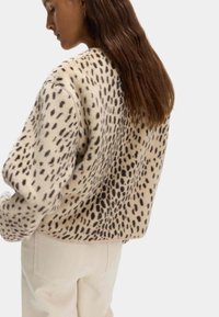 Přetáhnutý krémový svetr s černými leopardími skvrnami, měkká textura, spadlá ramena a žebrovaný lem; ve spojení s béžovými kalhotami.