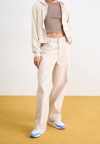 Světlé béžové krátké mikinové triko s kapucí, hnědý těsný top, béžové volné džíny a bílé tenisky s modrými akcenty na žlutém pozadí.