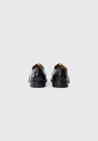 Une paire de chaussures de cérémonie en cuir noir vues de dos sur un fond blanc uni.