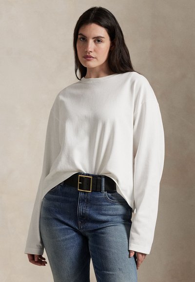 Kvinde med langt mørkt hår iført en løs hvid langærmet skjorte stoppet ned i blå jeans med sort bælte mod en neutral baggrund.