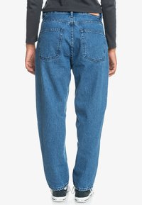 Heled sinised denim teksad, millel on lõdvestatud lõige, kaks tagumist taskut ja õmmeldud vöökoht, koos mustade tossudega.