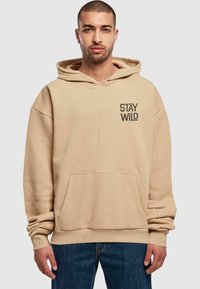 Bežs kapučinš ar kangaroo kabatu, apdrukāts ar melnu grafiku "STAY WILD", kam ir brīvs piegriezums un ribota apkakle un apakša.