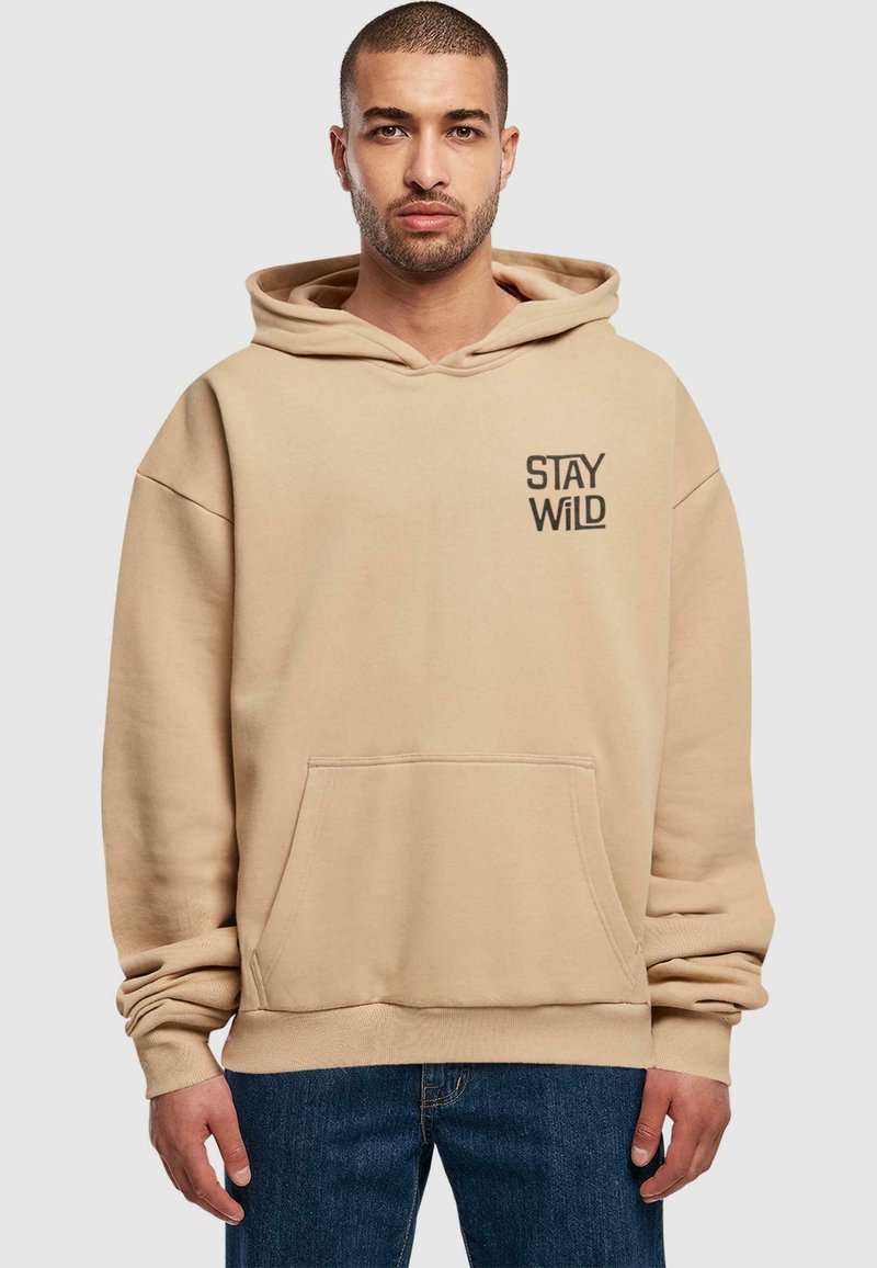 Bežs kapučinš ar kangaroo kabatu, apdrukāts ar melnu grafiku "STAY WILD", kam ir brīvs piegriezums un ribota apkakle un apakša.