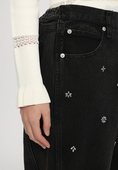 Black denim jeans with white flower embroidery and silver hardware details. The cuff features a textured white fabric with a ruffled edge.