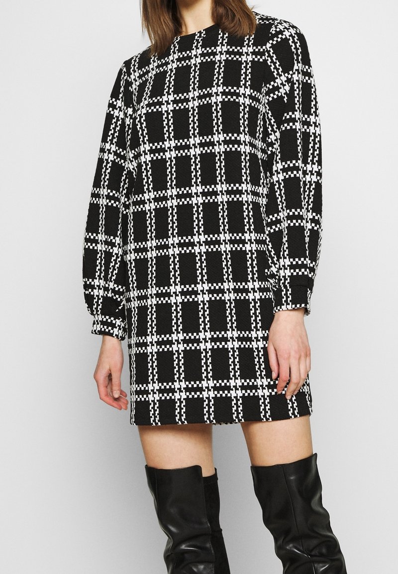 Femme portant une robe à manches longues à carreaux noir et blanc associée à des bottes en cuir noires montantes sur un fond uni.
