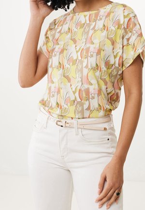 Femme portant un chemisier à manches courtes aux motifs abstraits pastel jaune, rose et blanc, rentré dans un pantalon taille haute blanc avec une ceinture beige.