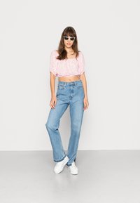 Blusa rosa cortada com padrão floral, jeans claros de cintura alta, e ténis brancos. Modelo em pé, a usar óculos de sol.