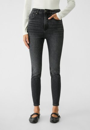 Persoon draagt zwarte high-waist skinny jeans met een witte top met lange mouwen en zwarte Mary Jane platte schoenen, staand tegen een effen achtergrond.