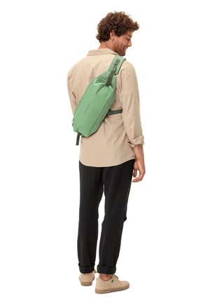 Sac en bandoulière vert en tissu texturé, doté de bretelles réglables et d'une forme épurée, porté sur une chemise beige et un pantalon noir.