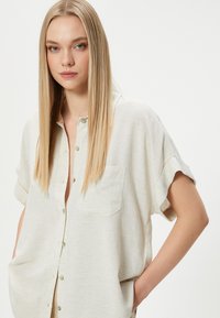 Camisa de manga corta en beige claro con parte delantera de botones, cuello y un bolsillo en el pecho. Hecha de una tela suave y texturizada.