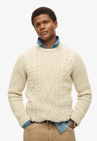 Cremefarbener Strickpullover mit strukturierten Kabelmustern und einem Diamantdesign, über einem blauen Jeanshemd getragen. Abgeschlossen mit gerippten Bündchen.