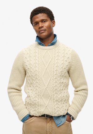 Pull en tricot couleur crème avec des motifs en relief et un design en losange, superposé sur une chemise en denim bleu. Fini avec des poignets côtelés.