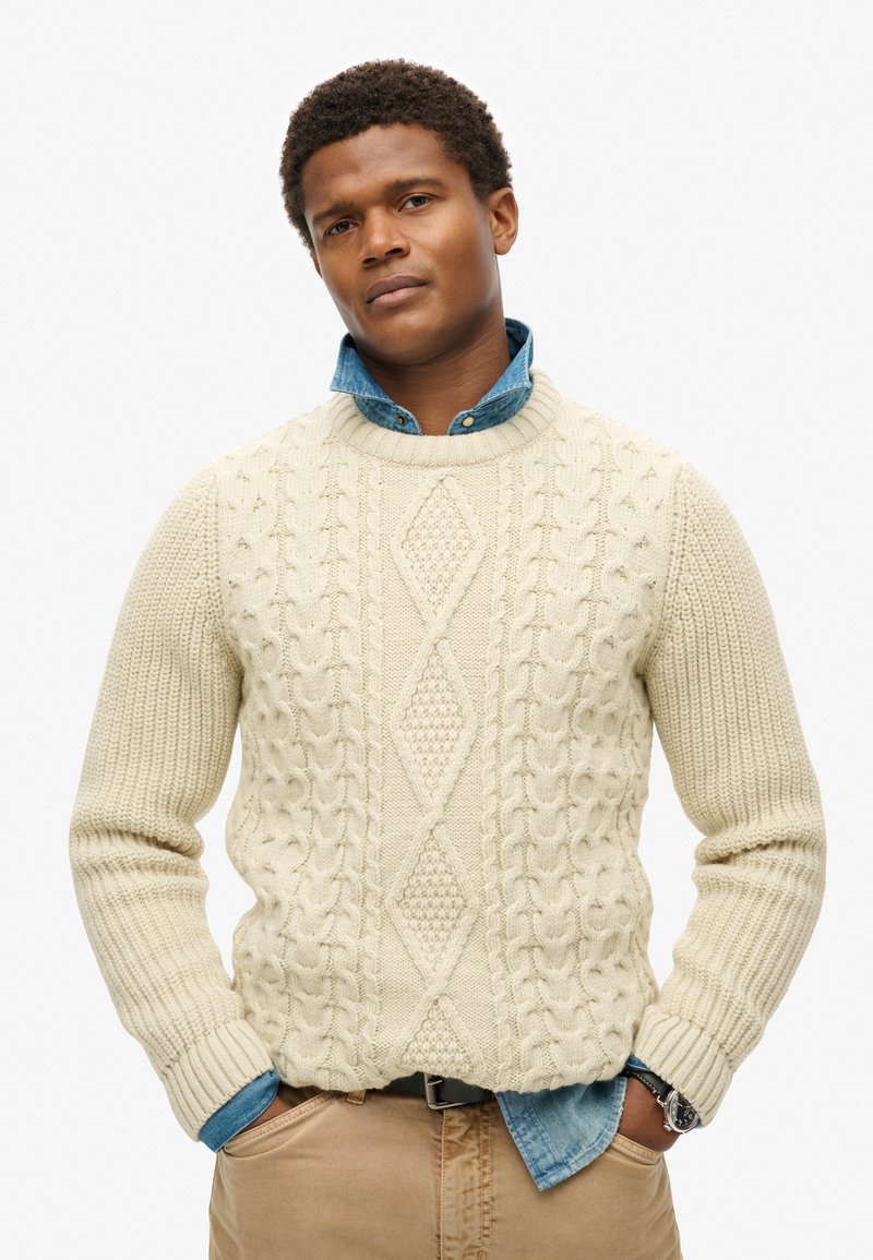 Cremefarbener Strickpullover mit strukturierten Kabelmustern und einem Diamantdesign, über einem blauen Jeanshemd getragen. Abgeschlossen mit gerippten Bündchen.
