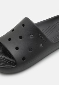 Svart sklisikret sandal laget av teksturert skummateriale med perforerte hull, glatt innside og preget merkevarelogo på stroppen.