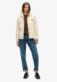 Cremefarvet lærredjakke med knaplukning, to brystlommer og rullede ærmer, parret med blå denimjeans og sorte sko.