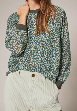 Nainen, jolla on pitkähihaista kuvioitua puseroa vihreillä kalvosimilla ja vaaleanbeiget corduroy-housut, käsi lepää lähellä kaulaa ja taskua.