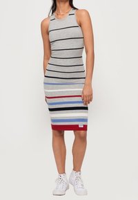 Nainen, jonka päällä on hihaton, polvipituinen harmaa mekko vaakasuorilla mustilla, punaisilla, sinisillä ja valkoisilla raidoilla, yhdistettynä valkoisiin korkeakorkoisiin tennareihin.