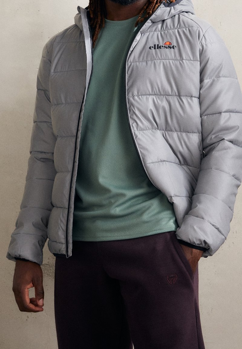 Homme portant une veste matelassée gris clair sur une chemise verte texturée et un pantalon foncé, debout devant un fond clair uni.