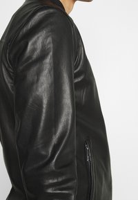 Svart läderjacka med en slät textur, åtsittande design och ribbad krage. Har en dragkedjeficka längs sidan.