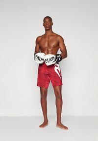 Σωματώδης άνδρας που στέκεται ξυπόλυτος, φορώντας κόκκινες σπορ σορτς και λευκά γάντια πυγμαχίας με το λογότυπο "VENUM", ποζάροντας μπροστά σε απλό φόντο.