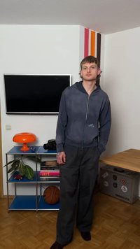 Ung mand i mørk lynlåstrøje og bukser står i et rum med et vægmonteret TV, en orange lampe, et kamera, en basketball og en stribet vægdekoration.