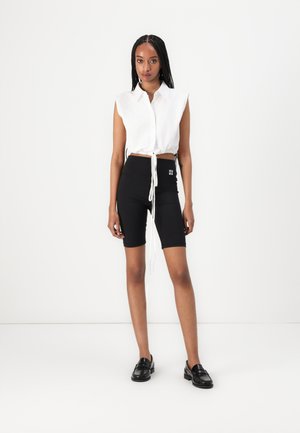 Camicia bianca corta con colletto e dettaglio cravatta, abbinata a pantaloni corti neri da ciclismo con logo e mocassini neri. Design semplice e aderente.