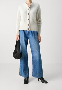 White wool cardigan with a v-neck and six brown buttons, paired with wide-leg light blue denim jeans and black heeled ankle boots. Black woven bag.