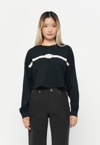 T-shirt noir à manches longues avec une bande blanche horizontale et un logo circulaire "ROXY". Assorti à un pantalon taille haute gris foncé.