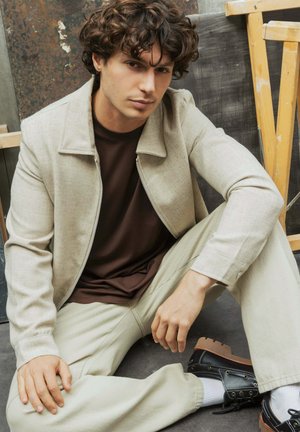 Jeune homme aux cheveux bouclés assis par terre, portant une veste beige, une chemise marron, un pantalon clair et des chaussures noires sur fond industriel.