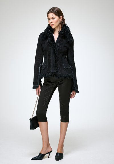 Femme en veste noire à col en fourrure, pantalon noir court et brillant, talons aiguilles noirs, tenant un petit sac noir avec chaîne dorée.