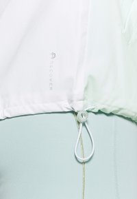 Biały dół kurtki z regulacją na sznurek, na jasnoniebieskich legginsach z pionowym szwem i złotymi zdobieniami.