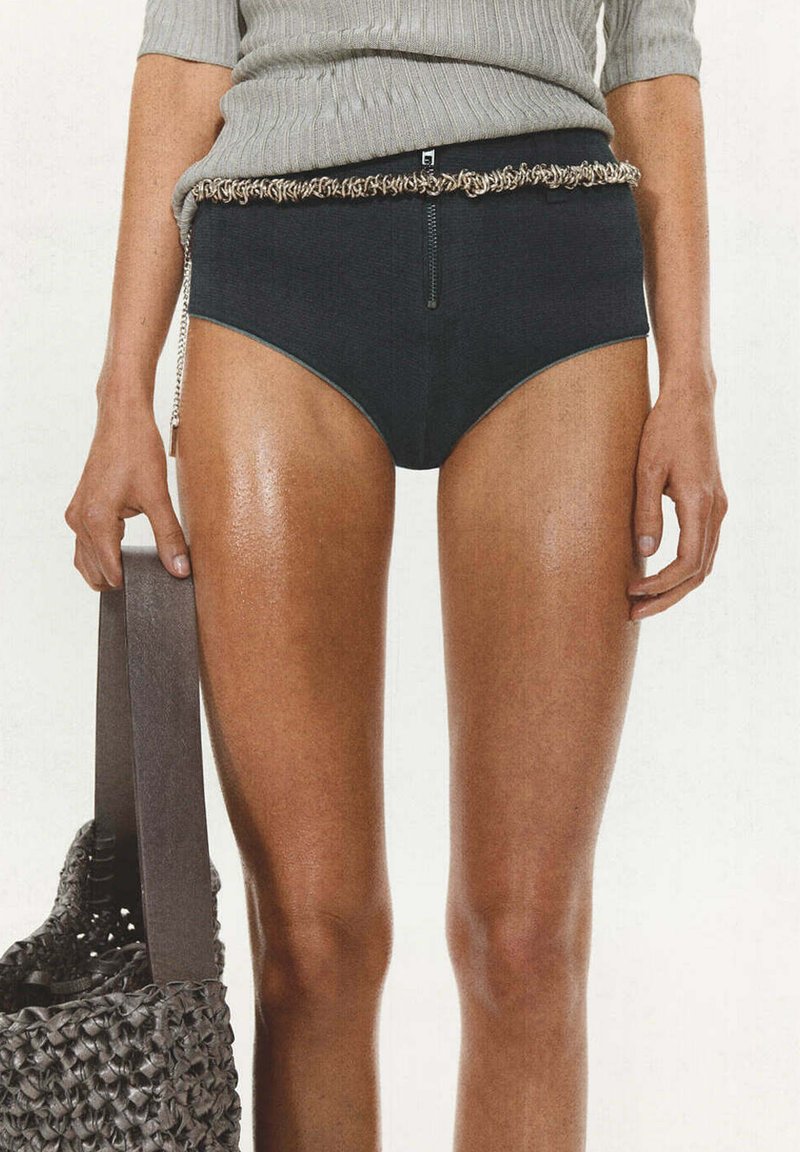 Personne portant une culotte noire taille haute avec ceinture chaîne, top gris côtelé, tenant un sac noir tissé à côté d’elle.
