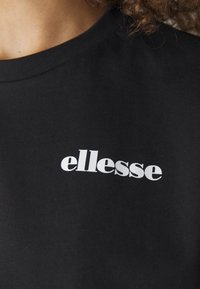 Juodas medvilnės marškinėliai su apvaliu iškirpimu ir baltu "ellesse" logotipu, spausdintu kairėje krūtinės pusėje. Lygi tekstūra.