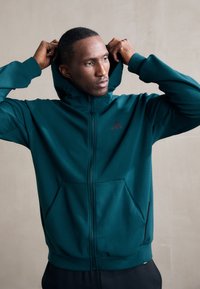 Teal zip-up hoodie with a front pocket, adjustable hood, and black logo on the chest. Made from smooth, lightweight fabric.
