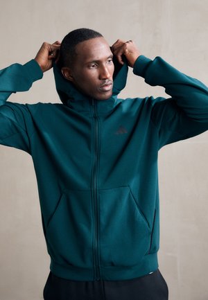 Teal zip-up hoodie med en frontlomme, justerbar hætte og sort logo på brystet. Lavet af glat, letstof.