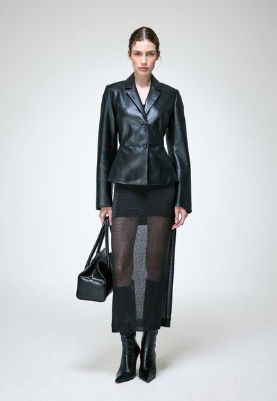 Femme portant une veste en cuir noire près du corps, une jupe longue noire transparente, des bottes noires et tenant un sac à main noir, debout devant un fond uni.