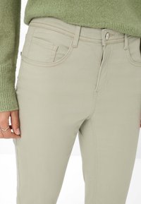 Primo piano di una persona che indossa pantaloni beige chiaro e un maglione lavorato a maglia verde, con tasche frontali cucite e chiusura con bottone.