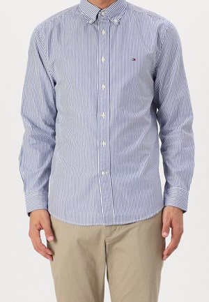 Uomo che indossa una camicia a bottoni a righe verticali blu e bianche con pantaloni beige, in piedi contro uno sfondo semplice.