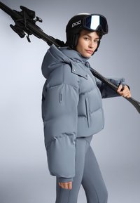 Svijetlosiva puffer jakna s kapuljačom, koja se ističe velikim kvadratnim dijelovima i prednjim zatvaranjem na dugmadi. Kombinira se s elegantnim usklađenim odijelom i ski opremom.