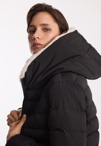 Czarna puchowa kurtka z wysokim kołnierzem, z miękkim beżowym futrem wewnątrz. Projekt zawiera pikowane sekcje i zamek błyskawiczny z przodu.