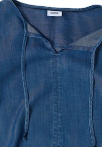 Blouse en chambray bleu avec un décolleté en V, deux liens à l'avant et une étiquette de marque "CECIL" au niveau du col.