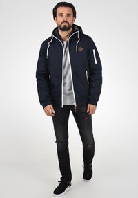 Marineblauwe pufferjack met trekkoorden, witte ritsaccenten, frontzakken, bruine logo-opprint, grijs shirt, zwarte versleten jeans en zwarte schoenen.