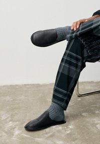 Black leather slip-on shoe worn with a gray ribbed sock. The individual is seated, displaying gray and black plaid patterned pants.