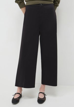 Schwarze weit geschnittene Hose aus glattem Stoff mit einem Knopfverschluss vorne und seitlichen Taschen. Getragen mit schwarzen flachen Schuhen.