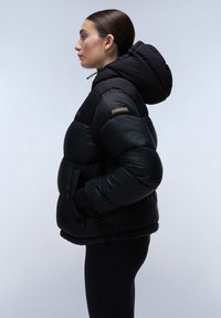 Giacca a vento nera con cappuccio, caratterizzata da una finitura lucida, polsini elasticizzati e una patch con logo sul petto. Le tasche laterali sono integrate nel design.