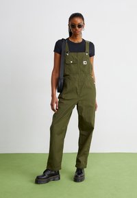 Olive green overalls with adjustable straps, front pockets, and metal hardware, paired with a black t-shirt and chunky black boots.