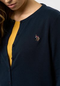 Cardigan tricotat navy cu decolteu rotund, cu butoane în față și un mic design brodat pe piept. Sub cardigan, se vede un top galben.