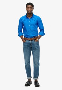 Blauwe button-up shirt, spijkerbroek, bruine riem, zwarte loafers. Shirt heeft opgerolde mouwen en de jeans zijn bij de enkels opgerold.