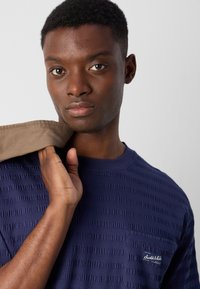 T-shirt texturé bleu marine avec un col rond et une petite poche poitrine. Le tissu présente un motif en rib et comporte une étiquette de logo sur la poche.