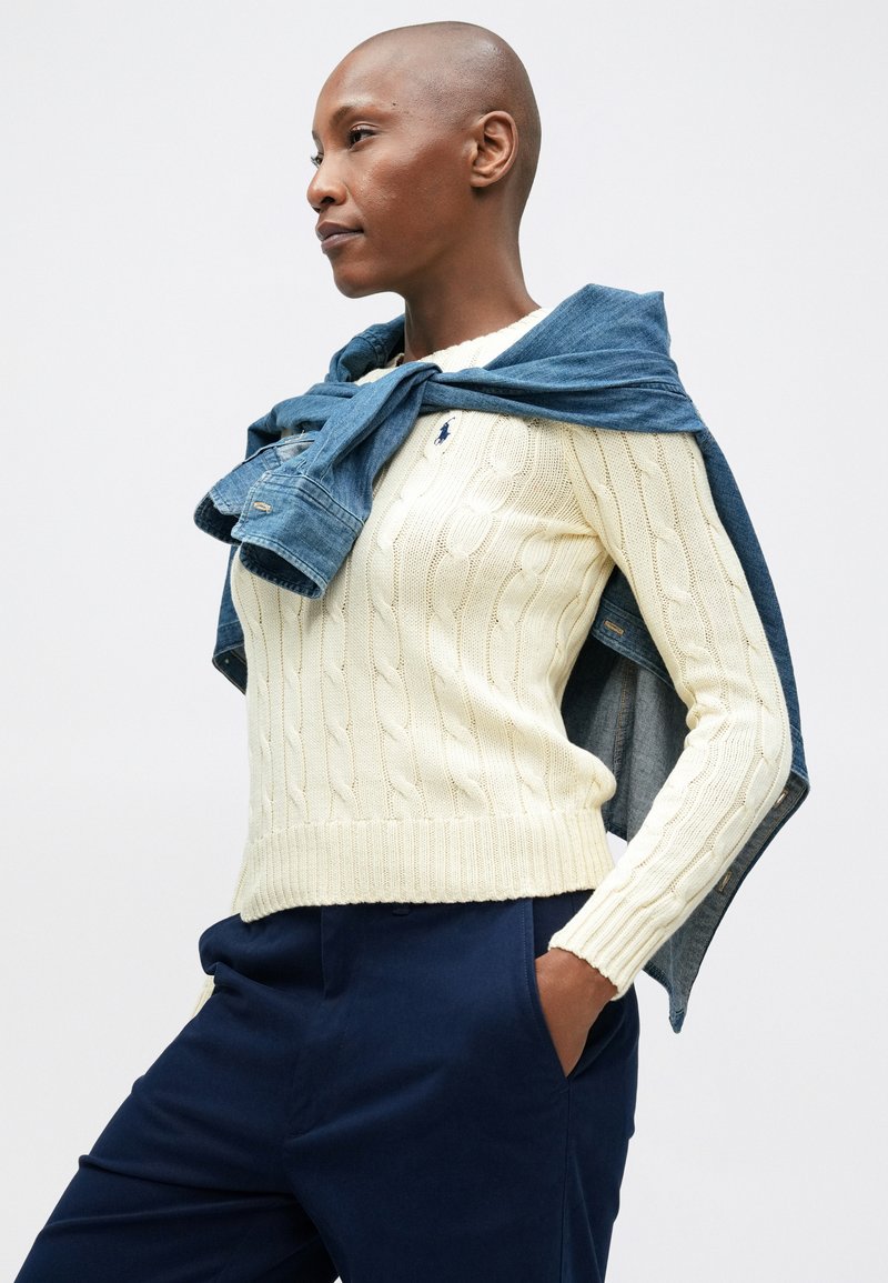 Pull en maille torsadée crème avec ourlet et poignets côtelés ; veste en denim nouée au cou ; pantalon bleu marine ; texture lisse, design ajusté.